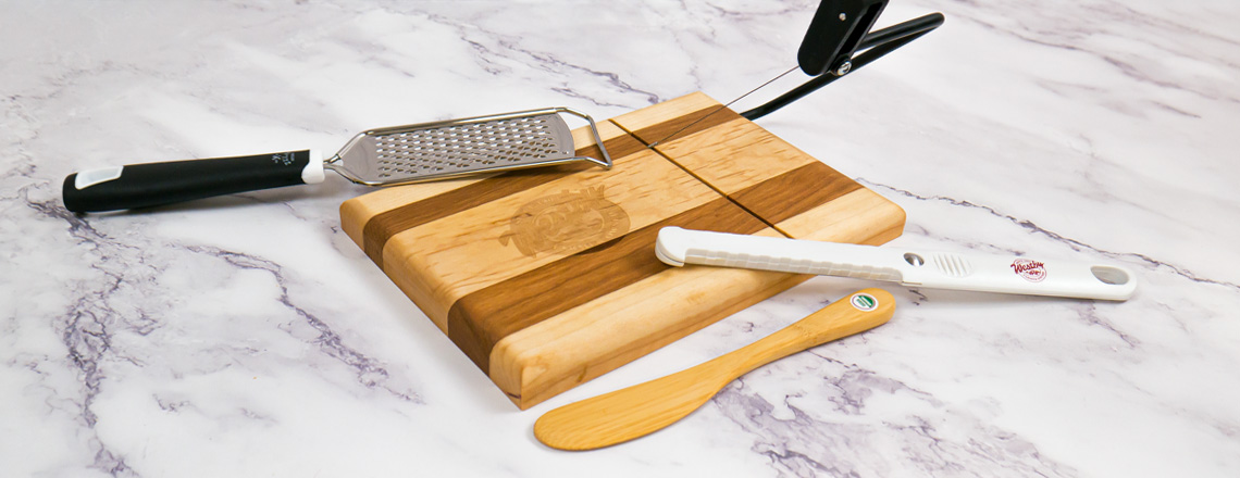 Assortment of Westby Cooperative Creamery kitchen gadgets - wooden cheese knife, wire cheese slicer, cheese grater, and cheese slicer and cutting board combo.