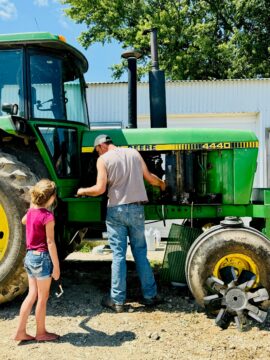 A father working on his broken down tractor while his daughter hands him tools.